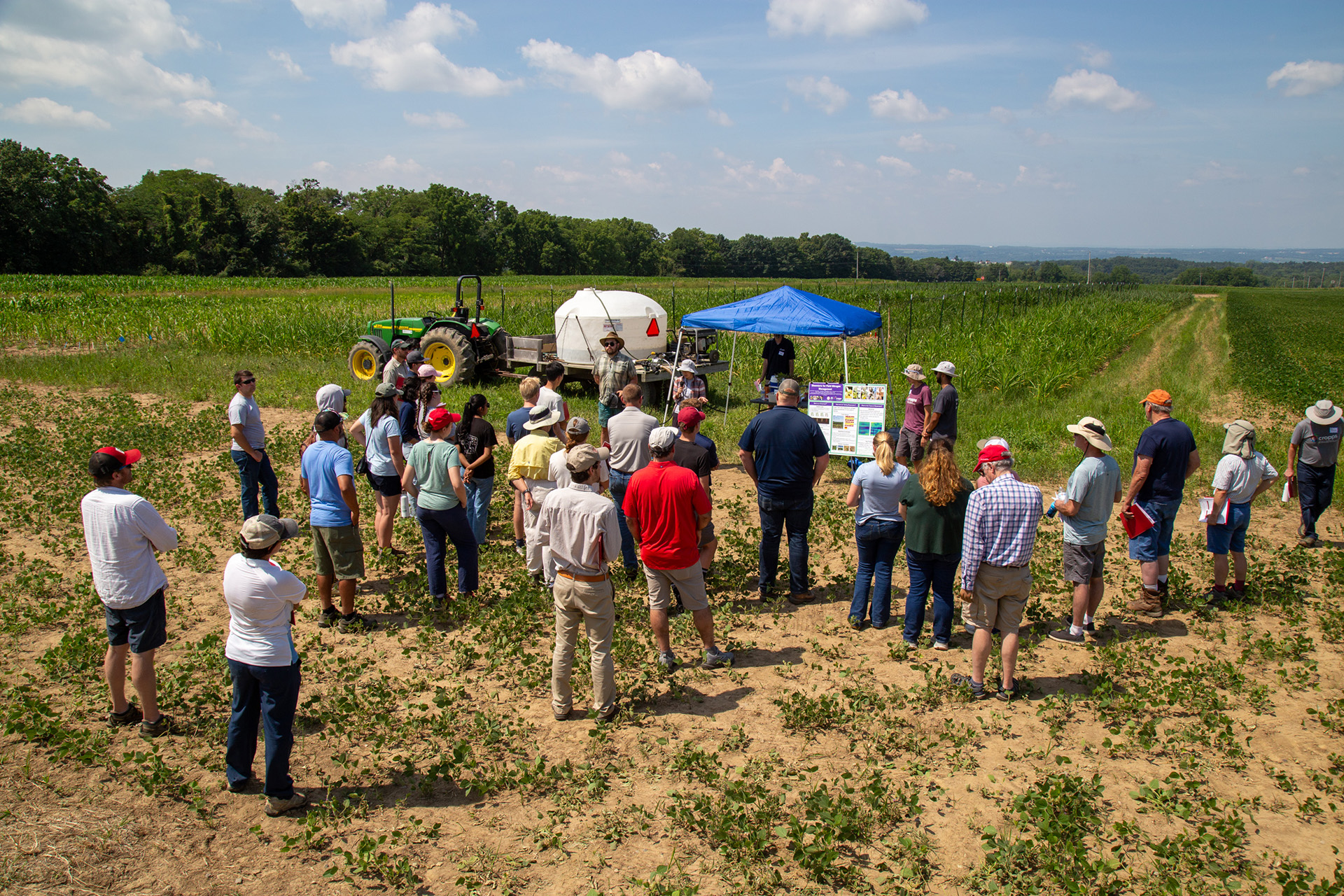 a large group of people stand in a cornfield listenning to a person explain the experiments happening in the field