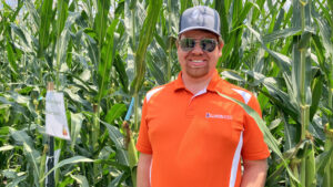 a man in an orange university of illinois urbana champaign shirt stands in front of a stand of corn with a gray baseball hat