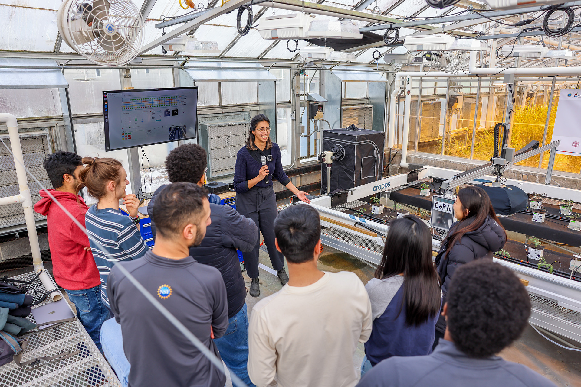 a woman speaks to a crowd gathered in front of a computer screen in a greenhouse